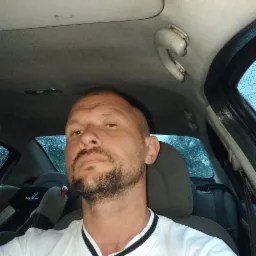 Mężczyzna z brodą w białej koszuli siedzący w samochodzie, widoczne krople deszczu na szybie