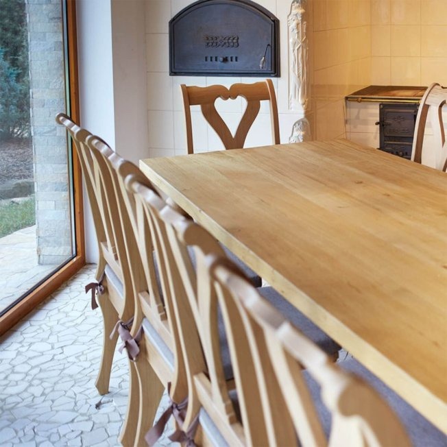 Jasny, drewniany stół jadalny z krzesłami o ażurowych oparciach w kształcie serc, ustawiony w pomieszczeniu z piecem kaflowym i oknem na ogród.