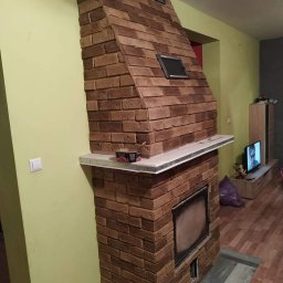 Swistak BUD - Murowany kominek z cegły klinkierowej w salonie, z metalowymi kratkami wentylacyjnymi i półką z blatem z jasnego kamienia, widoczny fragment telewizora w tle.