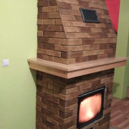 Swistak BUD - Wykończony kominek z cegły wewnątrz pomieszczenia z drewnianą półką i palącym się ogniem.