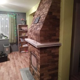 Swistak BUD - Murowany kominek z cegły klinkierowej z półką, widoczne kratki wentylacyjne, szara ściana w tle z oknem i zasłonami oraz drewniana podłoga.