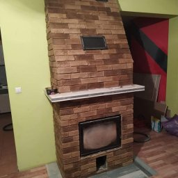 Swistak BUD - Murowany kominek z cegły klinkierowej w trakcie budowy, z widocznymi otworami wentylacyjnymi i półką, na podłodze częściowo ułożone panele.