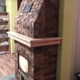 Murowany kominek z cegły z drewnianą półką, widoczne palenisko z płomieniami, kratki wentylacyjne i fragment wnętrza z regałem na książki.