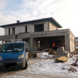 Swistak BUD - Niedokończony, dwukondygnacyjny dom z pustaków, z niebieskim vanem zaparkowanym przed nim na ośnieżonym terenie budowy, widoczny pracownik w pomarańczowej kamizelce wewnątrz budynku.