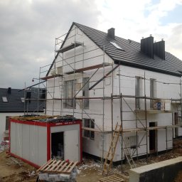 Swistak BUD - Dom jednorodzinny w trakcie ocieplania styropianem, widoczne rusztowania, kontener budowlany i materiały budowlane na palecie.