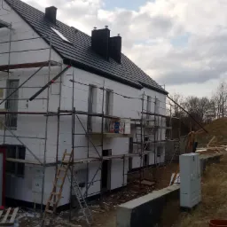 Dom w trakcie ocieplania styropianem, widoczne rusztowania, czarna dachówka i skrzynki gazowe na ścianie.