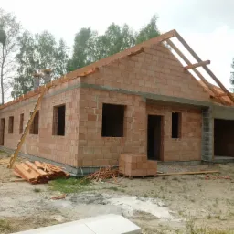 Niedokończony dom jednorodzinny z czerwonej cegły ceramicznej, widoczne drewniane elementy konstrukcji dachu, drabina oparta o ścianę, deski i cegły na placu budowy.