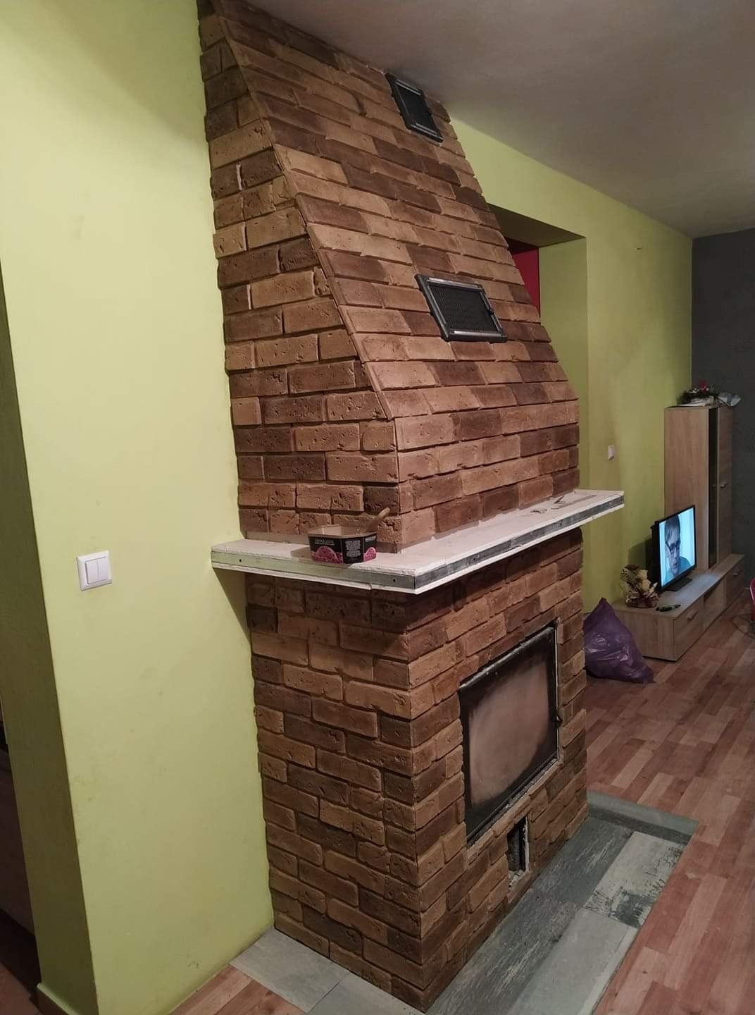 Murowany kominek z cegły klinkierowej w salonie, z metalowymi kratkami wentylacyjnymi i półką z blatem z jasnego kamienia, widoczny fragment telewizora w tle.