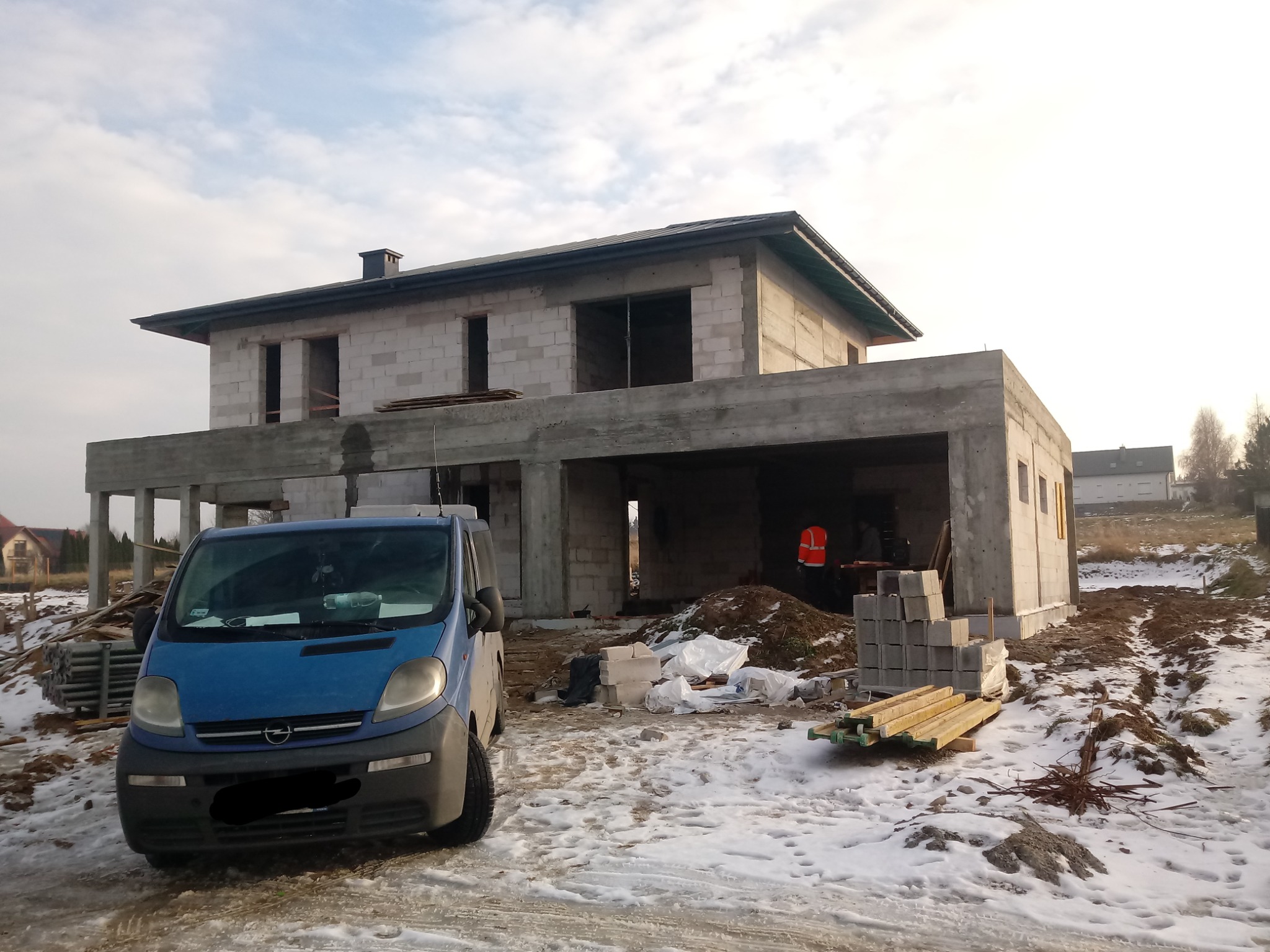 Niedokończony, dwukondygnacyjny dom z pustaków, z niebieskim vanem zaparkowanym przed nim na ośnieżonym terenie budowy, widoczny pracownik w pomarańczowej kamizelce wewnątrz budynku.