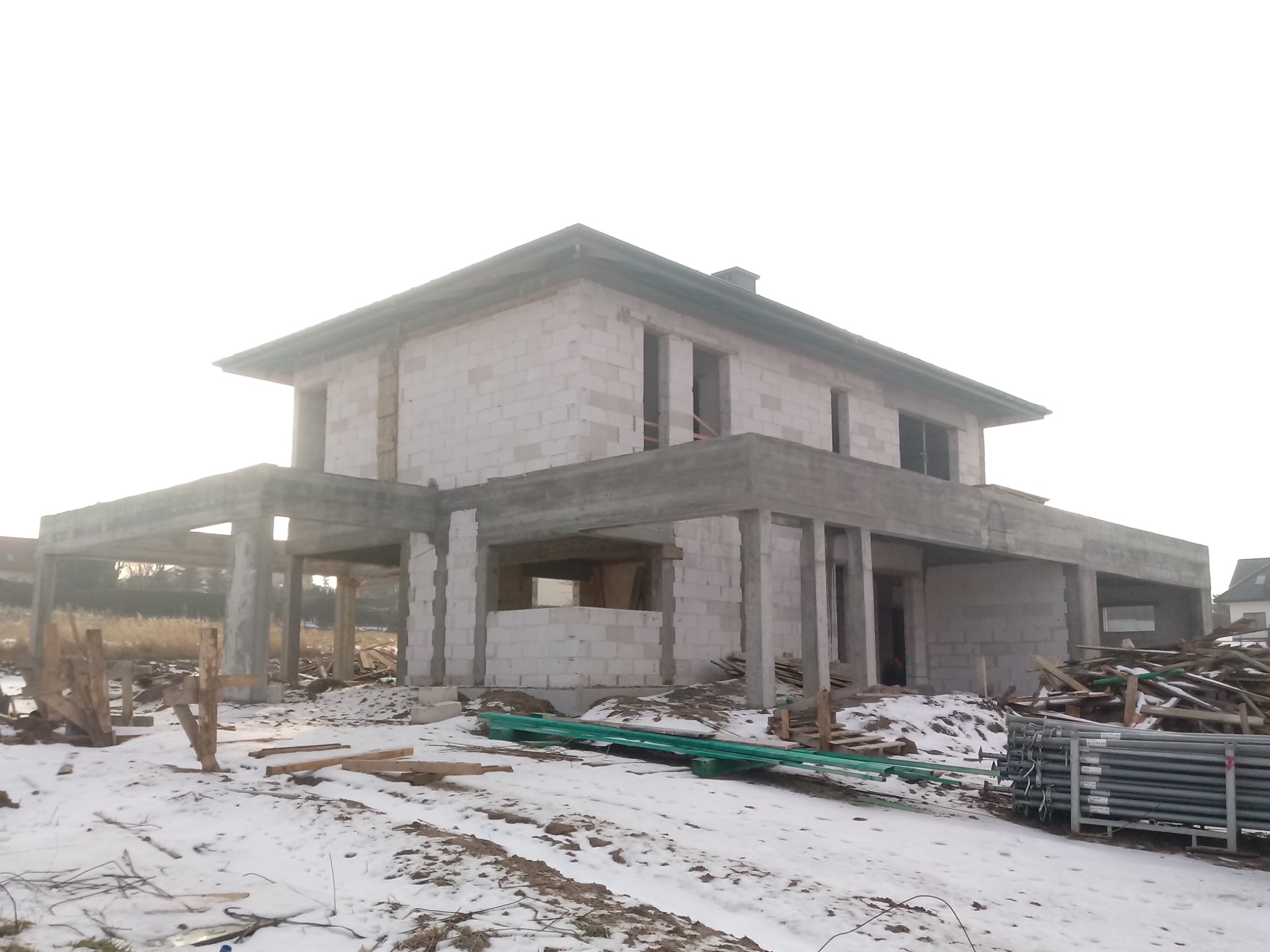 Surowy stan domu jednorodzinnego w trakcie budowy, ściany z betonu komórkowego, widoczne zbrojenia i podpory, teren wokół pokryty śniegiem.