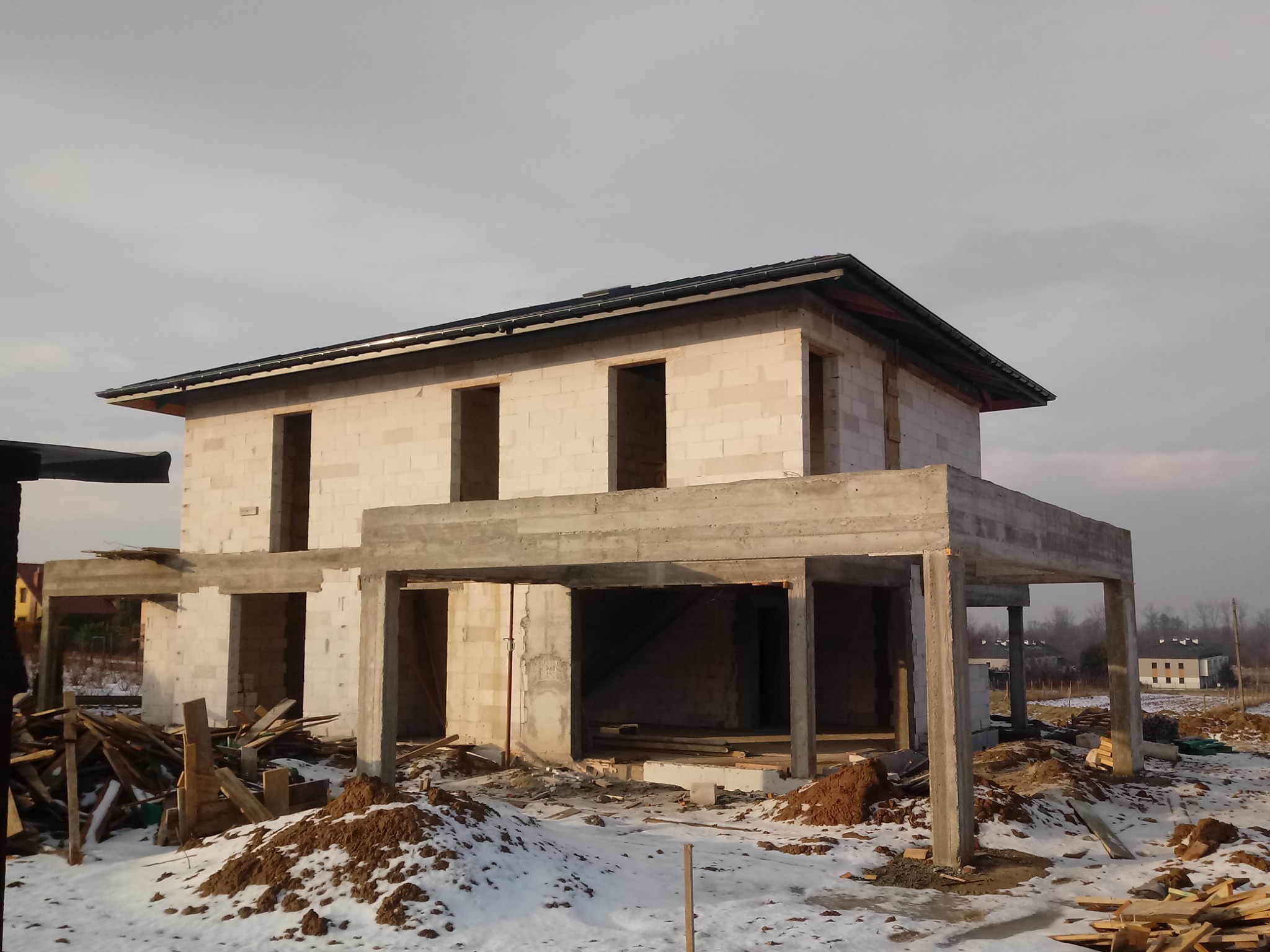 Surowy stan dwukondygnacyjnego domu z betonu komórkowego z widocznymi otworami okiennymi i betonową konstrukcją wsporczą, ziemia wokół pokryta resztkami śniegu.