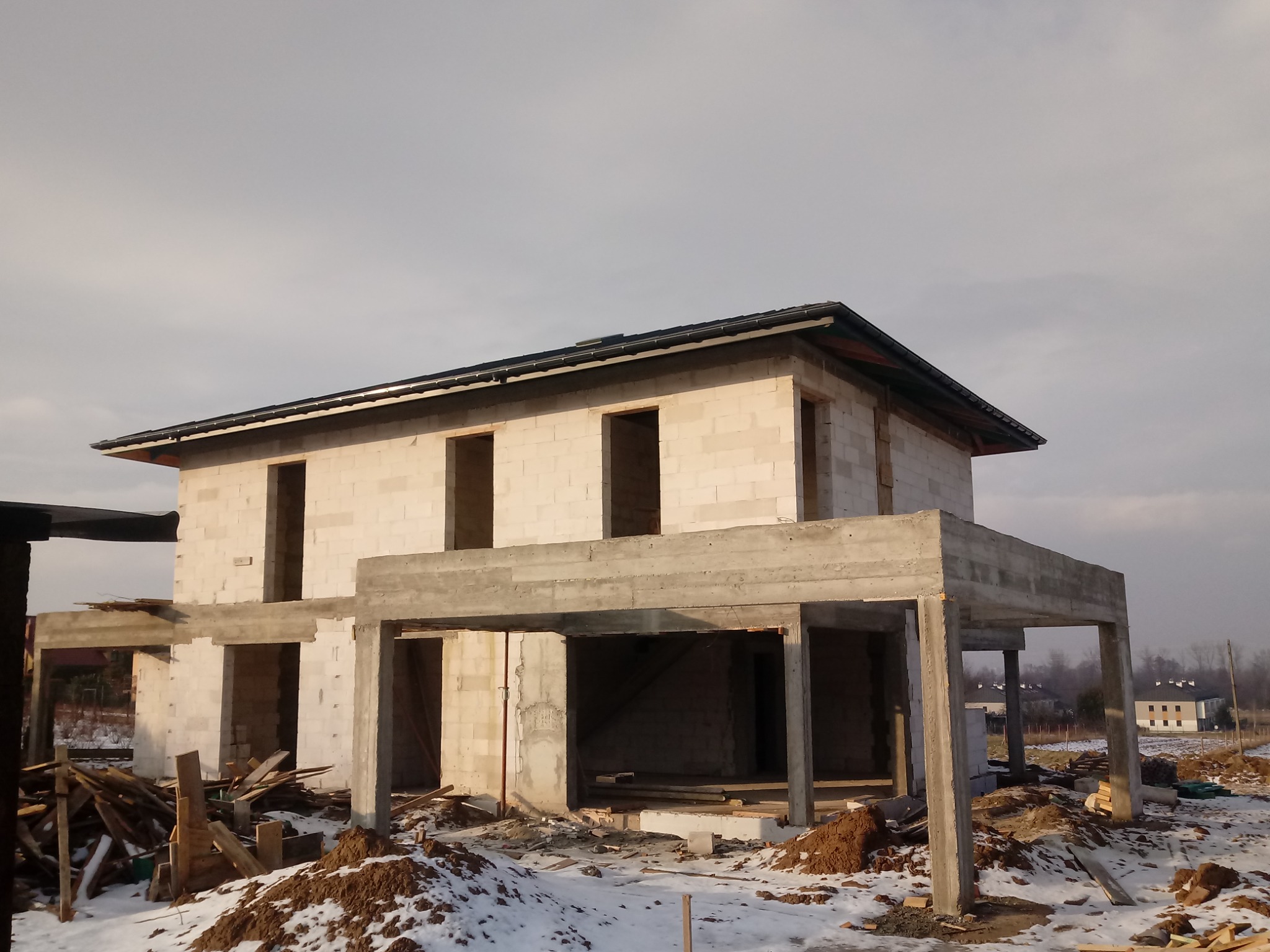 Surowy stan dwukondygnacyjnego domu z betonu komórkowego i surowego betonu, z widocznym zadaszeniem, na działce pokrytej śniegiem i ziemią, w tle zabudowania.