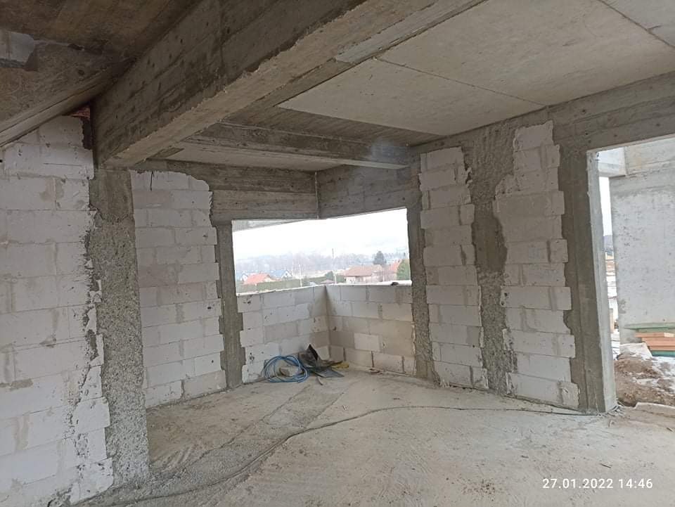 Surowe wnętrze poddasza w trakcie budowy: białe bloczki, betonowe belki stropowe, widok na zabudowę miejską w tle przez otwór okienny, kable i narzędzia na podłodze.