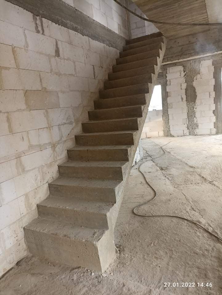 Surowe betonowe schody w trakcie budowy, białe bloczki ścian, widoczne przewody, wnętrze w stanie deweloperskim.