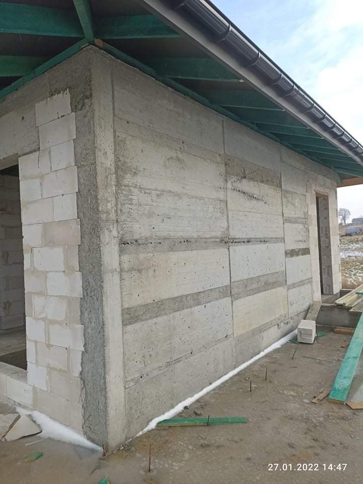 Surowa ściana zewnętrzna budynku w trakcie budowy, z widocznymi blokami z betonu komórkowego na rogu i poziomymi pasami szarego betonu, uszczelniona białą pianą, z zielonymi elementami konstrukcji...