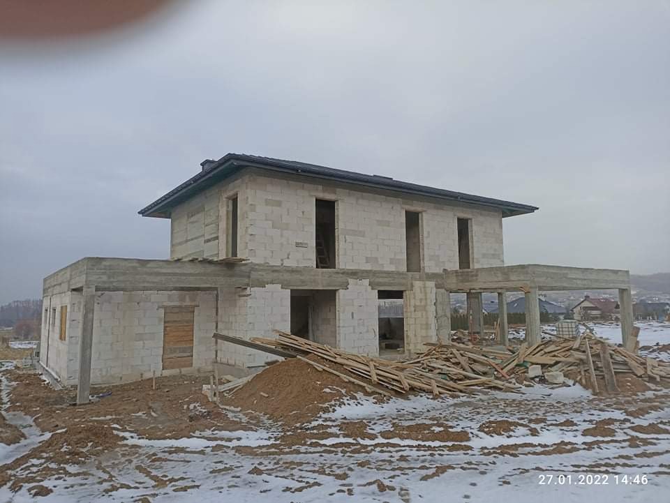 Surowy stan dwukondygnacyjnego domu z betonu komórkowego, widoczne otwarte przestrzenie okienne i drzwiowe, nieotynkowane ściany, sterta drewna na zasypanym śniegiem terenie budowy, data na dole...