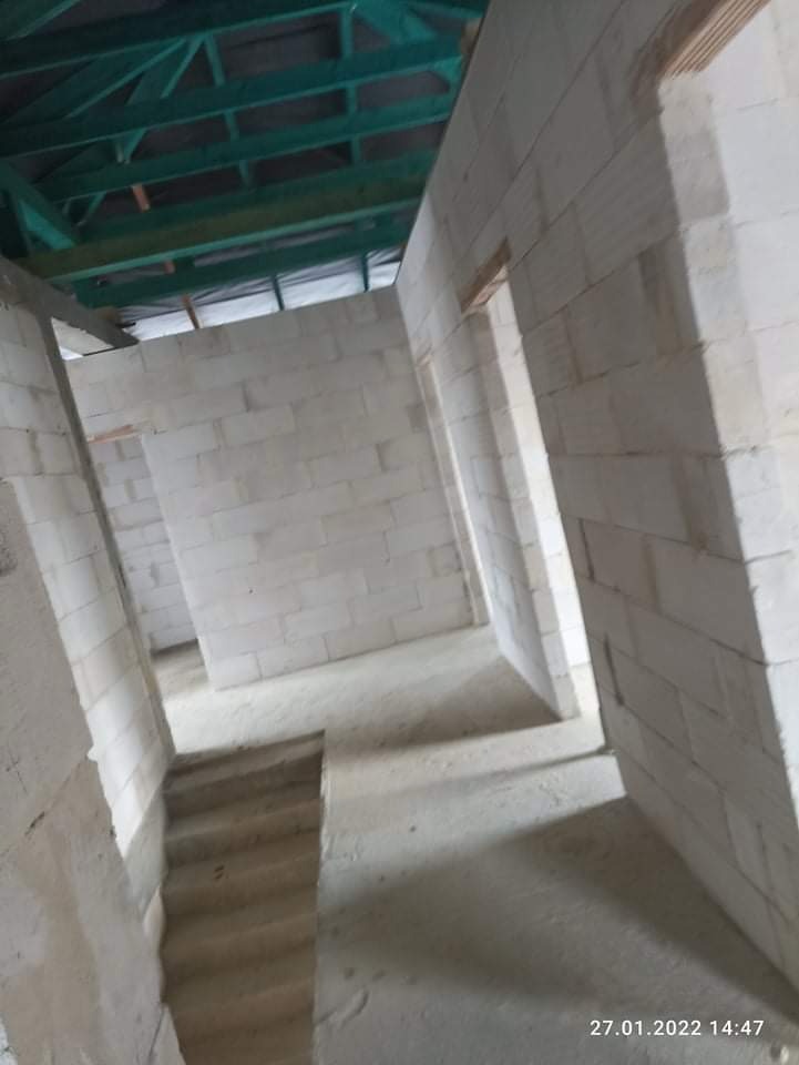 Surowe wnętrze domu w trakcie budowy: białe ściany z bloczków, betonowa podłoga i zielona konstrukcja dachu.