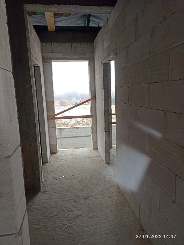 Wnętrze surowego budynku w trakcie budowy, widoczne ściany z bloczków betonowych, otwory okienne z drewnianymi rozpórkami, surowa podłoga i widok na zewnątrz na zabudowę jednorodzinną w oddali.