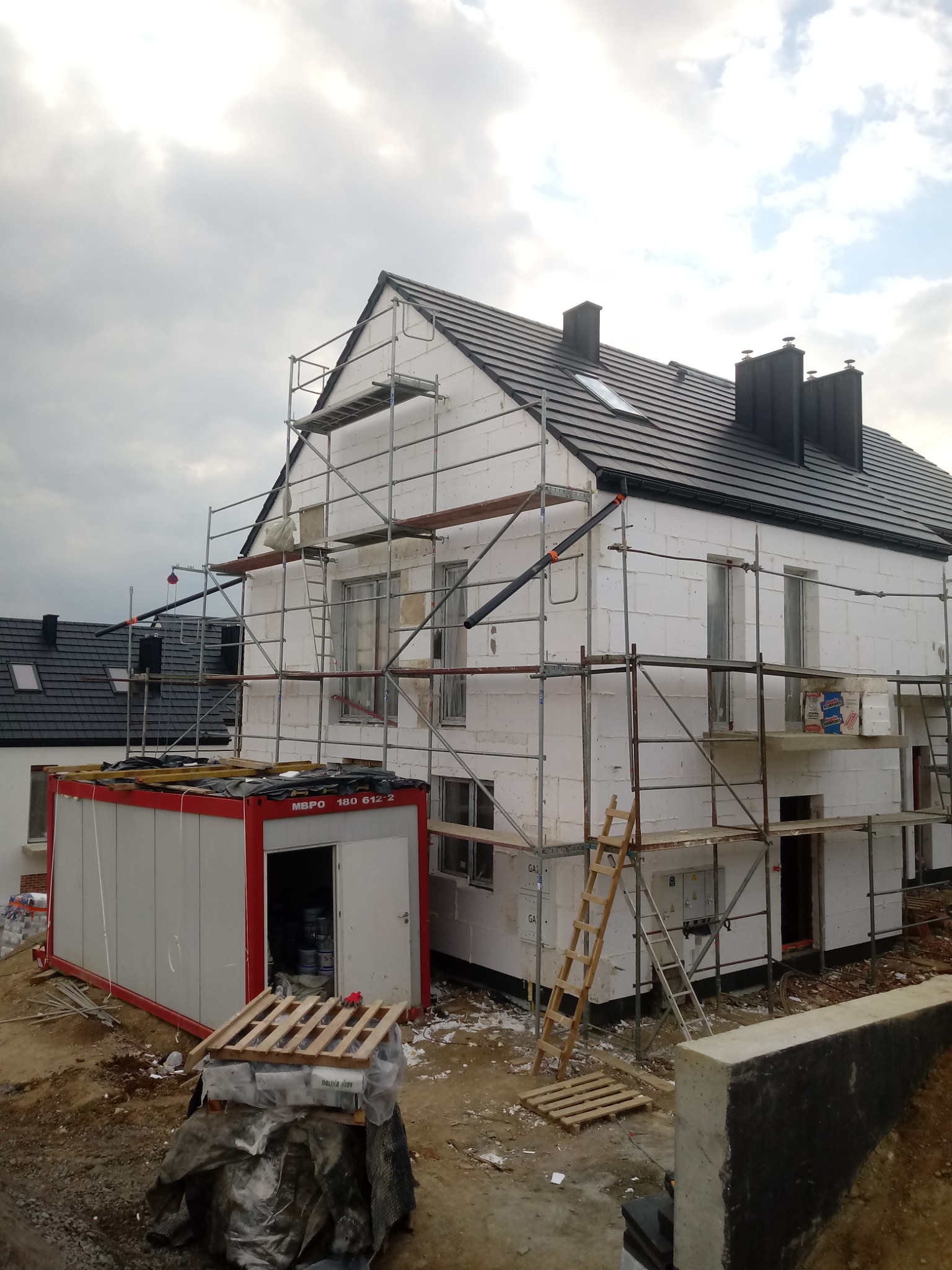 Dom jednorodzinny w trakcie ocieplania styropianem, widoczne rusztowania, kontener budowlany i materiały budowlane na palecie.