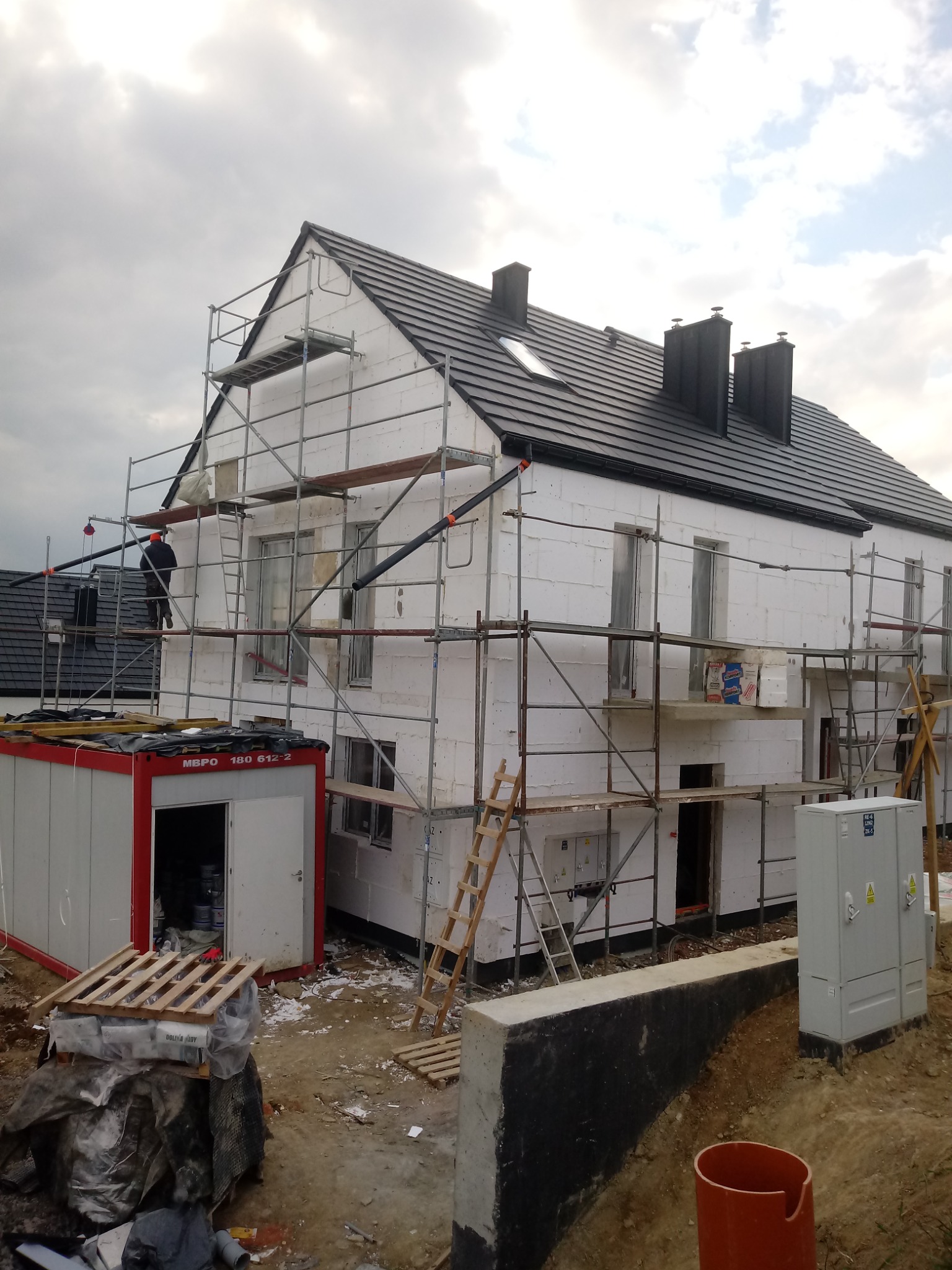 Dom w trakcie ocieplania styropianem, widoczne rusztowania, kontener budowlany i materiały izolacyjne na paletach.