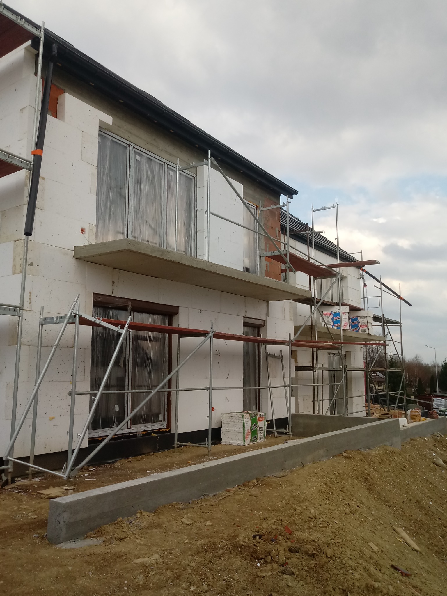 Dom w trakcie ocieplania styropianem, z widocznymi rusztowaniami, oknami zabezpieczonymi folią, oraz częściowo wykonaną podmurówką