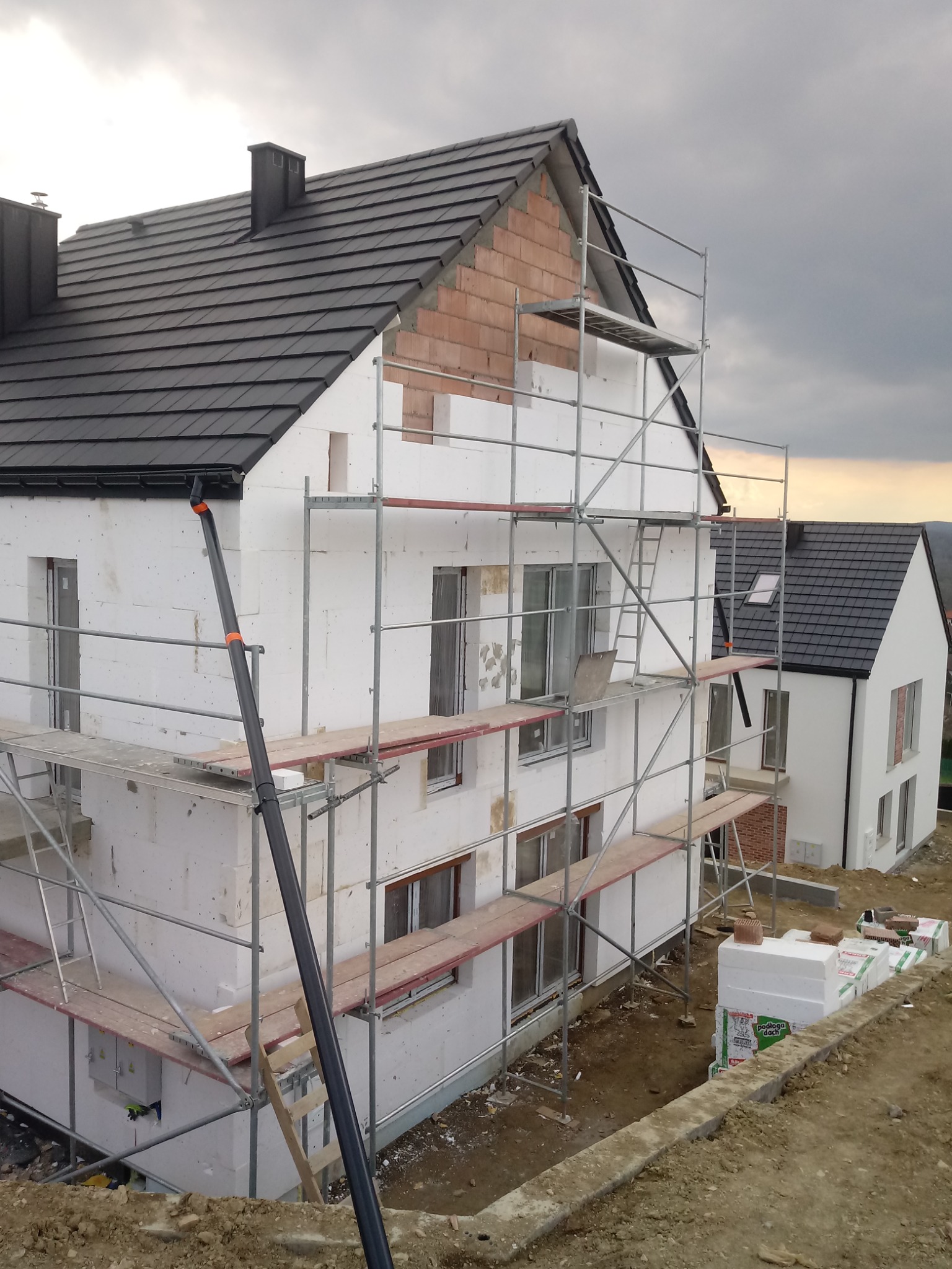 Dom w trakcie ocieplania styropianem, widoczne rusztowania, niezakończona elewacja i fragmenty muru z cegły, obok drugi dom z gotową elewacją.