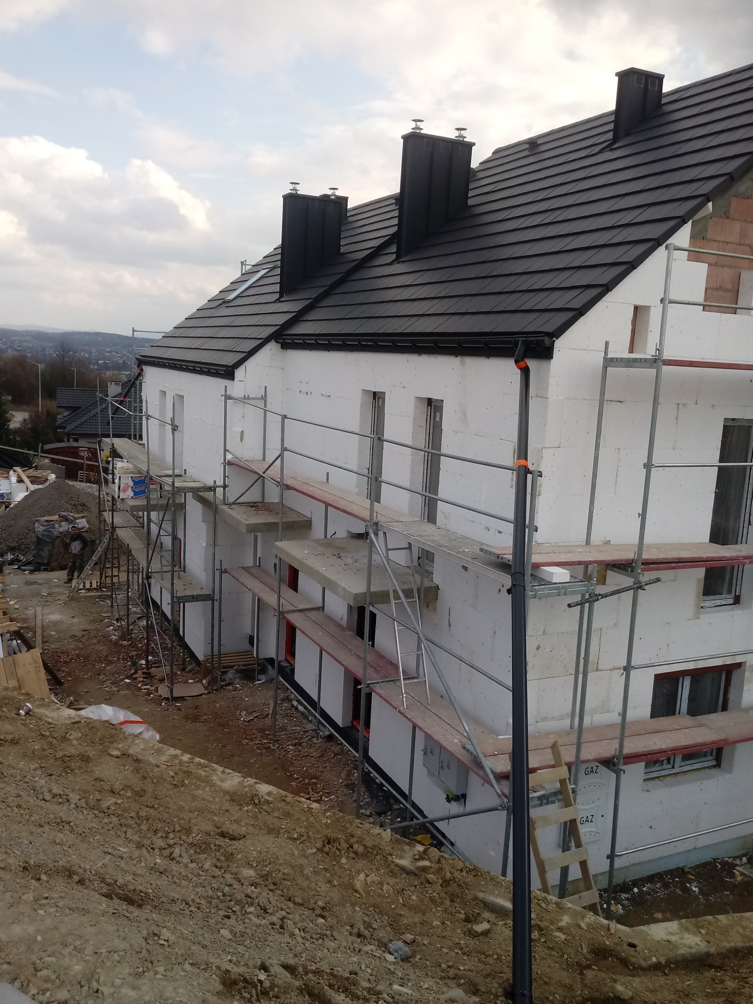 Dom jednorodzinny w trakcie ocieplania styropianem, widoczne rusztowania, czarna dachówka i kominy, teren wokół w stanie surowym.
