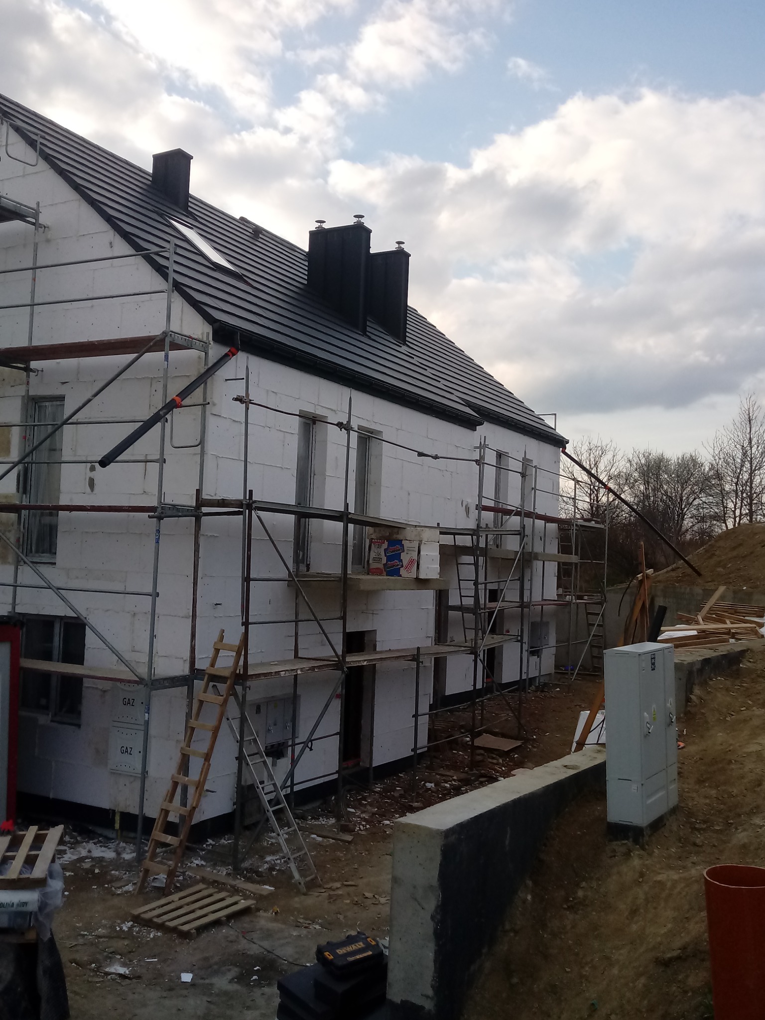 Dom w trakcie ocieplania styropianem, widoczne rusztowania, czarna dachówka i skrzynki gazowe na ścianie.