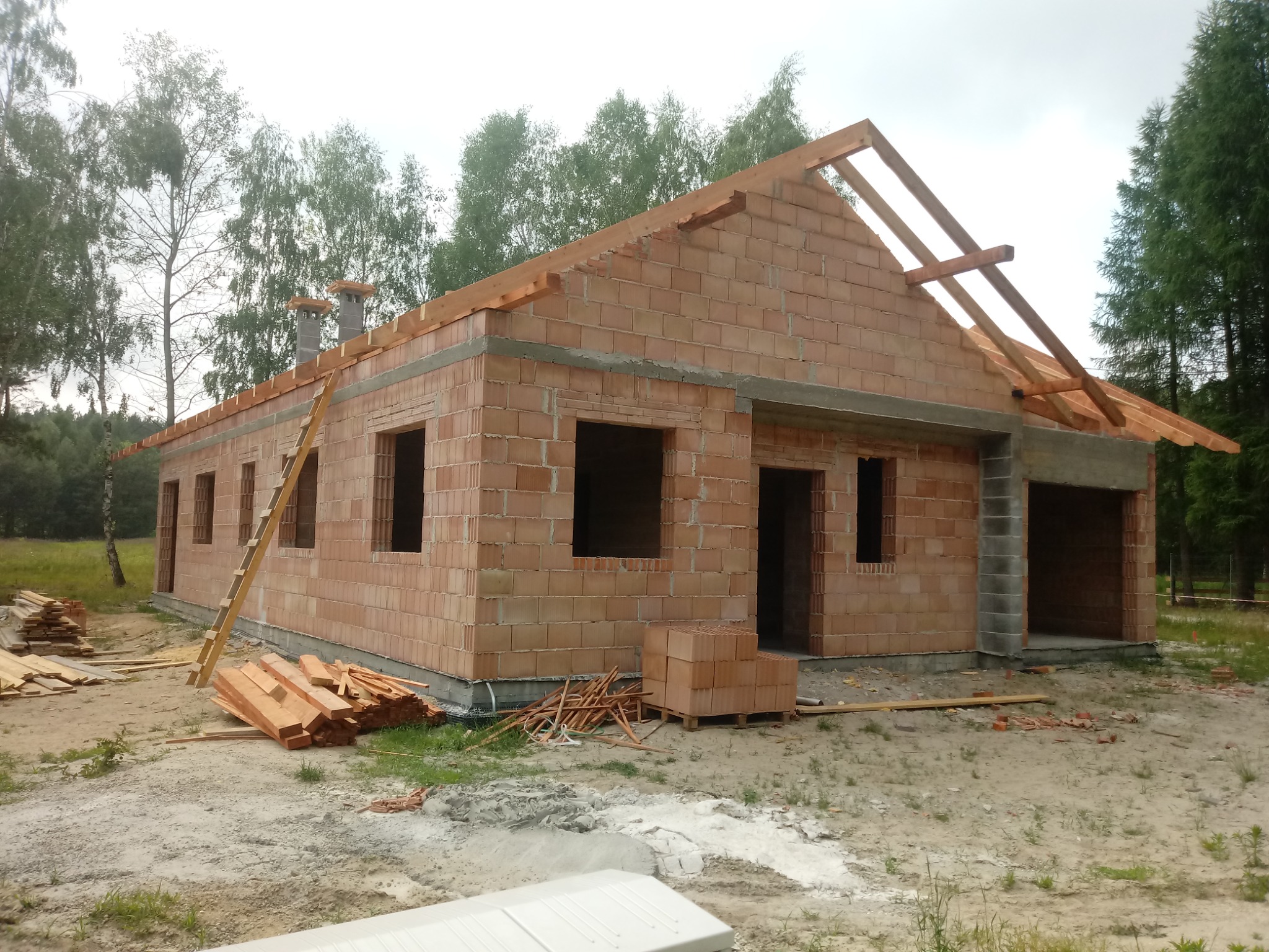 Niedokończony dom jednorodzinny z czerwonej cegły ceramicznej, widoczne drewniane elementy konstrukcji dachu, drabina oparta o ścianę, deski i cegły na placu budowy.