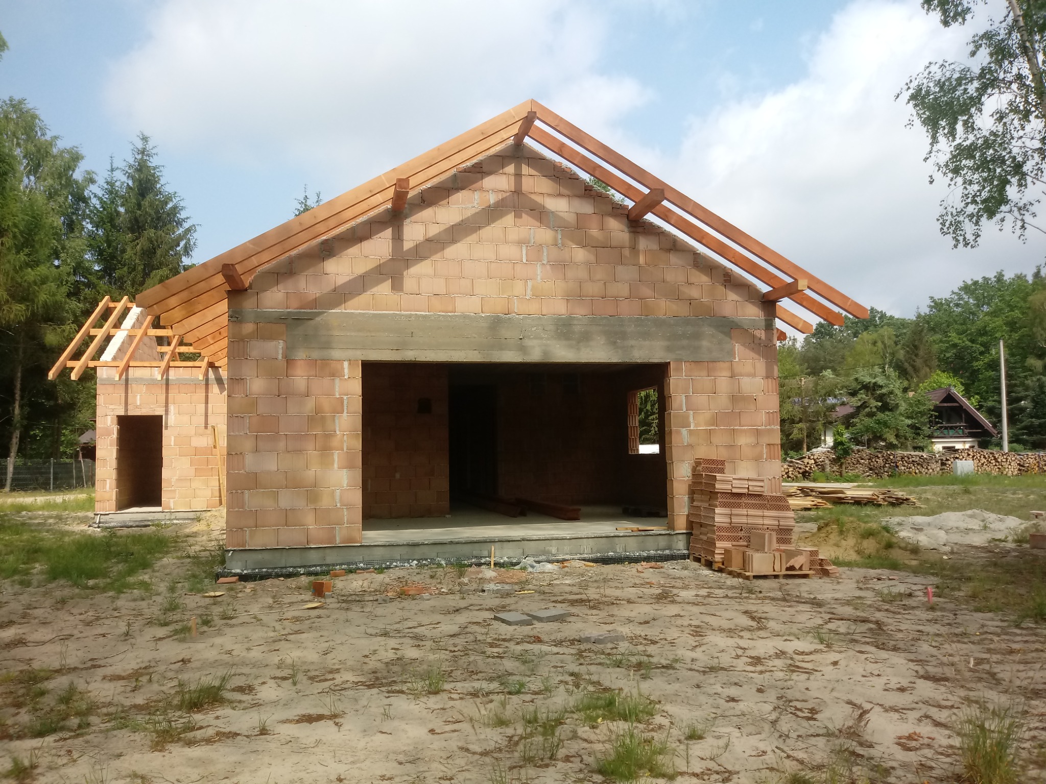 Budowa domu jednorodzinnego w stanie surowym otwartym: ściany z bloczków ceramicznych, drewniana konstrukcja dachu, widoczne materiały budowlane na posesji, otoczenie leśne.