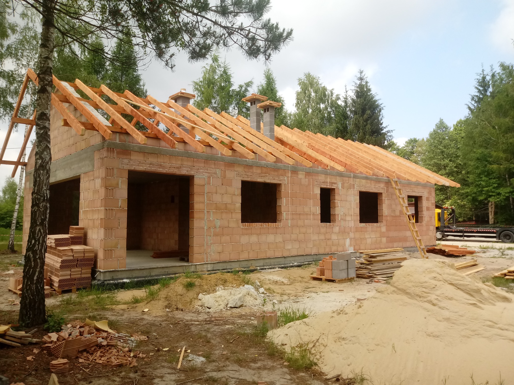 Surowy stan domu z czerwonej cegły z drewnianą konstrukcją dachu w trakcie budowy, widoczne kominy i drabina oparta o ścianę, otoczenie placu budowy z piaskiem i drzewami.