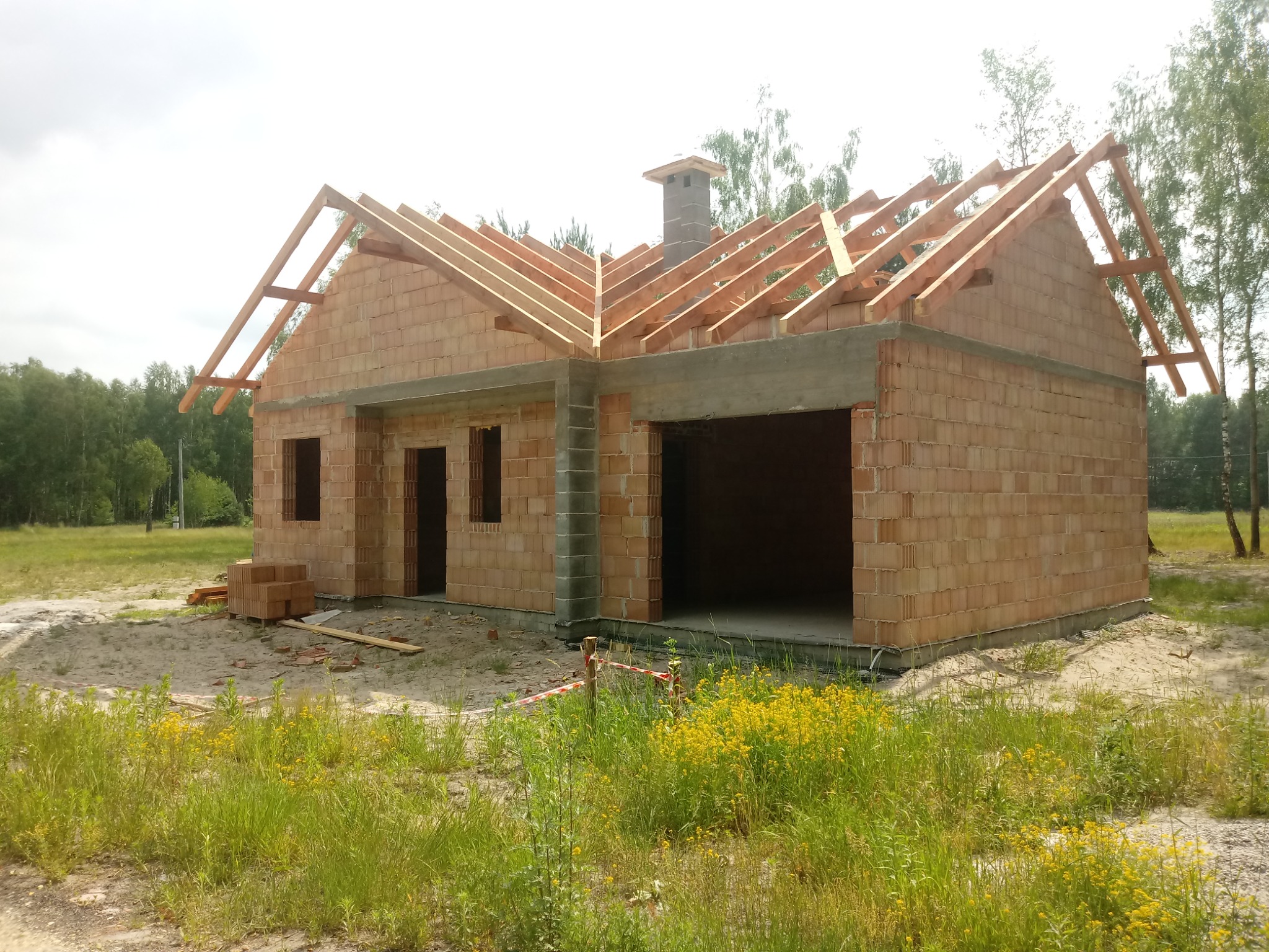 Surowy stan domu jednorodzinnego z cegły ceramicznej: widoczne ściany, otwory okienne i drzwiowe, drewniana konstrukcja dachu oraz komin. Na pierwszym planie wysoka trawa i żółte kwiaty.