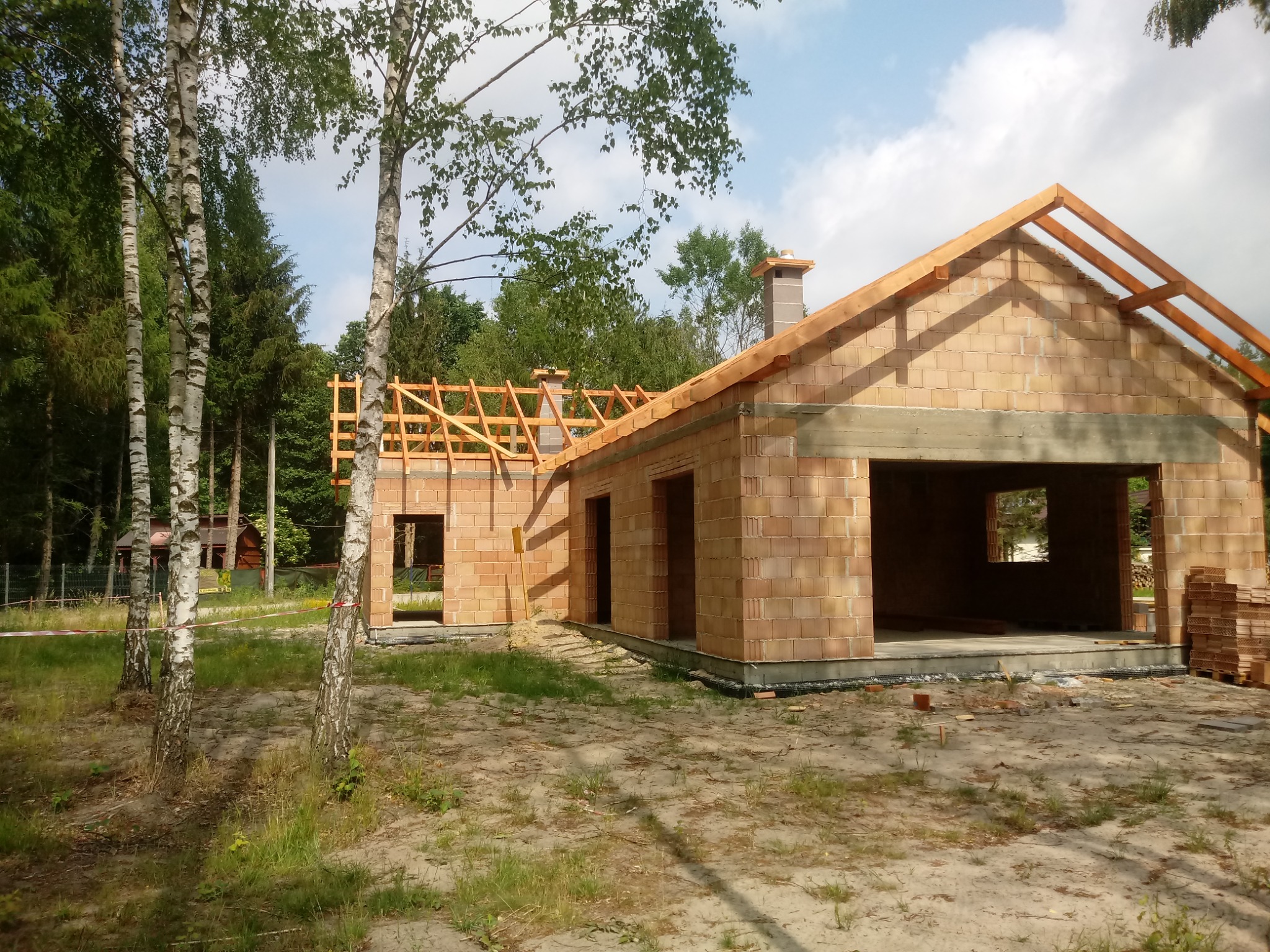 Budowa domu jednorodzinnego w stanie surowym otwartym, z widoczną konstrukcją dachu, murowanymi ścianami z pustaków ceramicznych, kominem i drewnianymi elementami więźby dachowej, na działce...