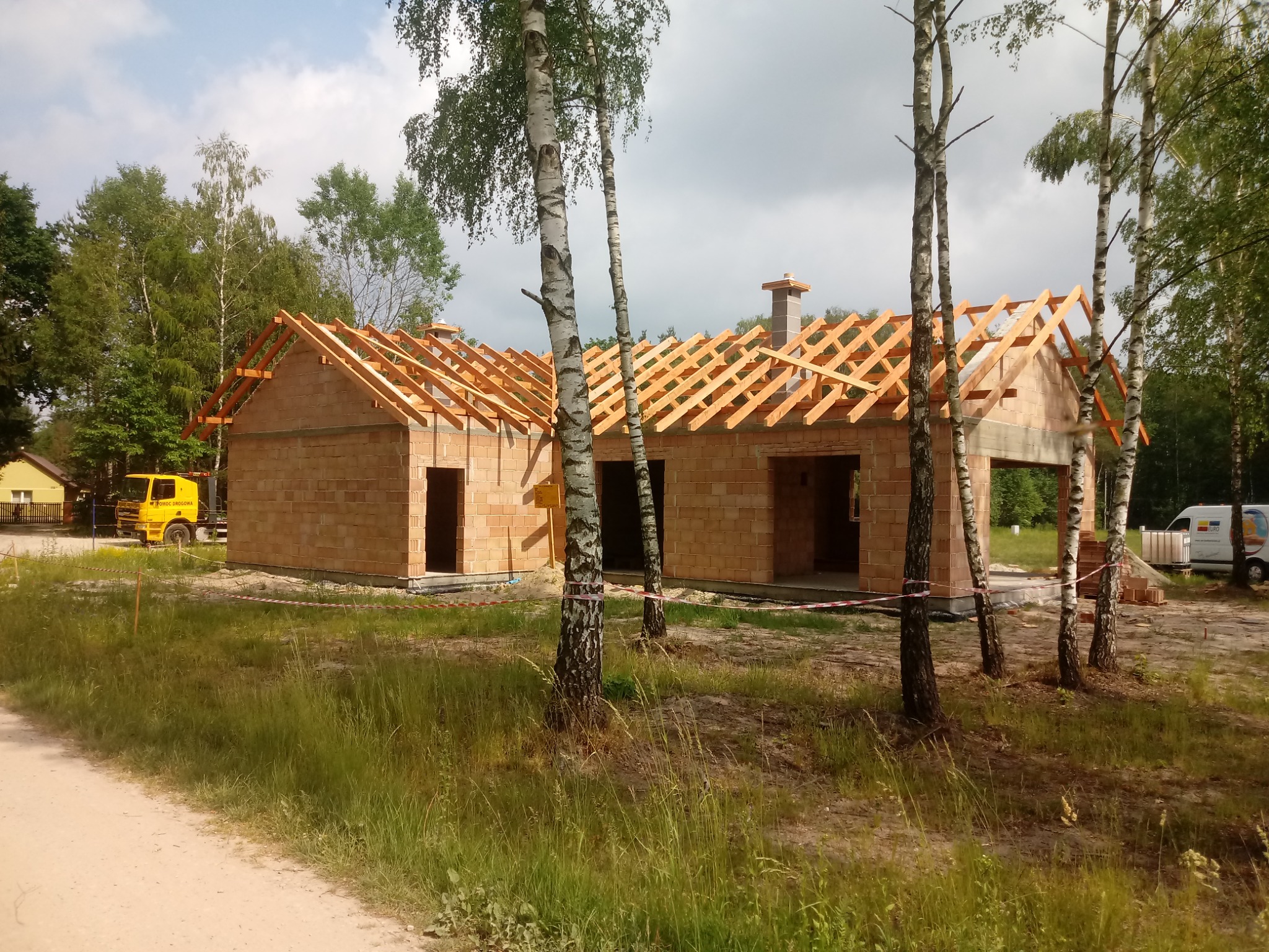 Surowy budynek z cegły z drewnianą konstrukcją dachu w trakcie budowy, widok z zewnątrz, otoczony młodymi brzozami i trawą, z żółtą ciężarówką w tle.