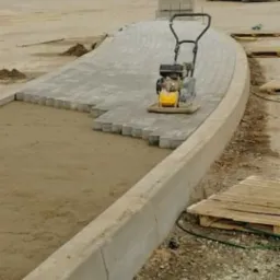 Etap prac: zagęszczarka wibrująca na ułożonej części ścieżki z kostki brukowej, widoczne przygotowane podłoże i obrzeża betonowe. W tle zaparkowany biały samochód dostawczy.