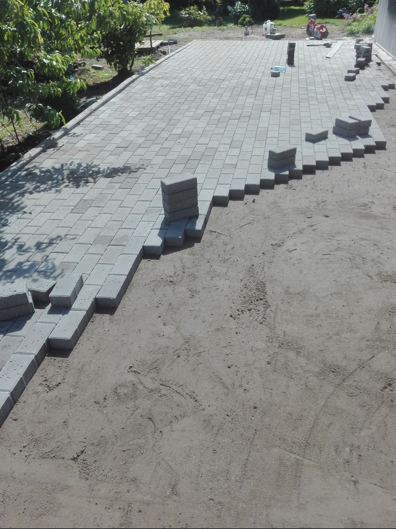 Układanie szarej kostki brukowej na podsypce piaskowej, widoczne narzędzia i częściowo ułożona nawierzchnia z regularnym wzorem. Praca w toku.