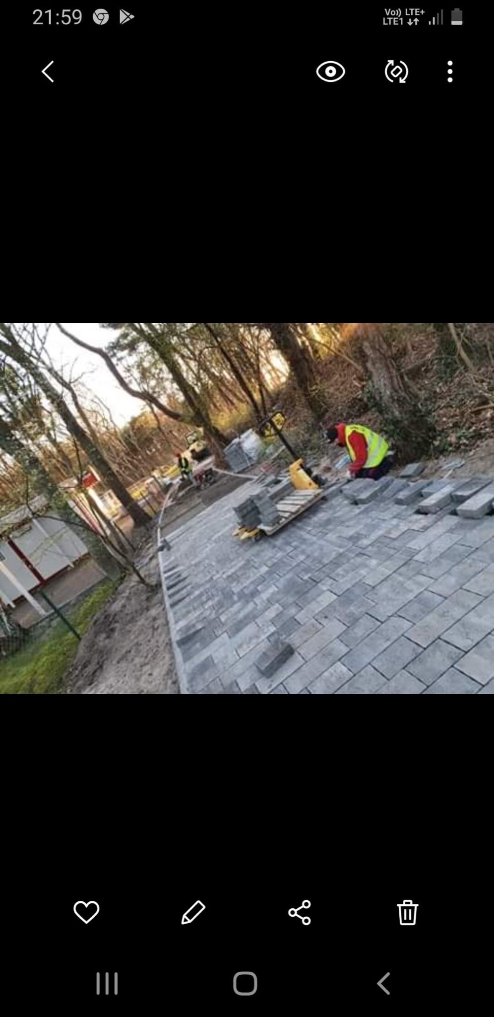 Prace brukarskie w Koszalinie: Układanie szarej kostki na posesji, widoczny pracownik w kamizelce odblaskowej i paleta z kostką oraz maszyna do zagęszczania.