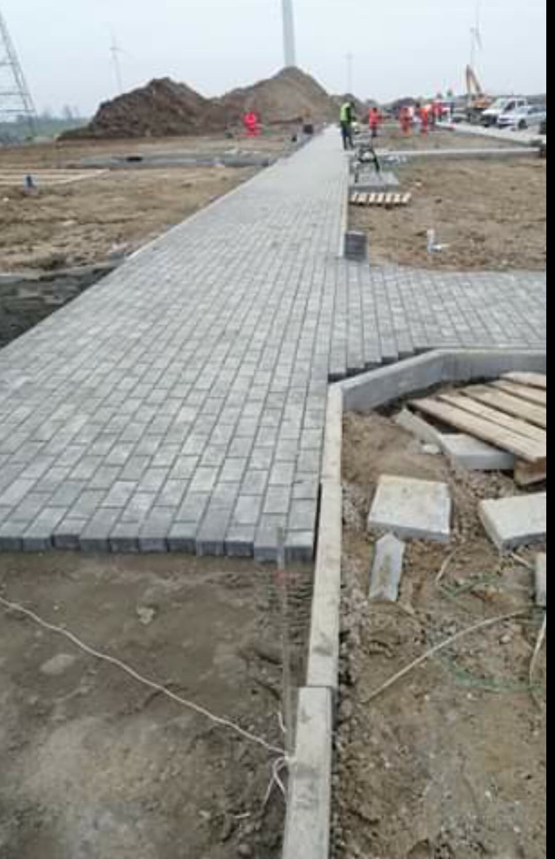 W trakcie układania szarej kostki brukowej na długim chodniku z widocznymi pracownikami w oddali i turbinami wiatrowymi w tle.