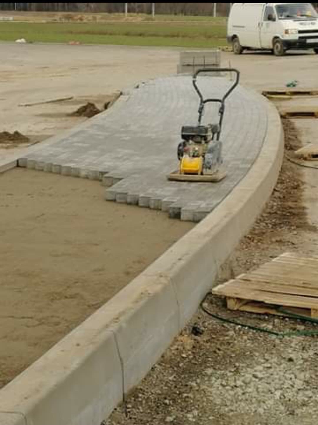 Etap prac: zagęszczarka wibrująca na ułożonej części ścieżki z kostki brukowej, widoczne przygotowane podłoże i obrzeża betonowe. W tle zaparkowany biały samochód dostawczy.
