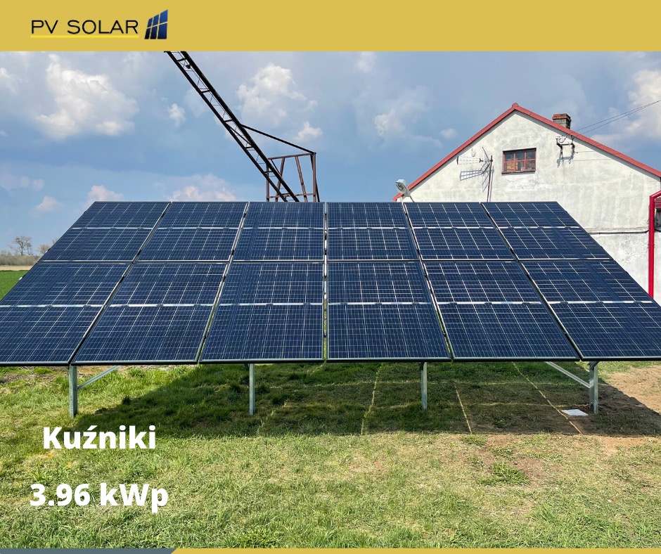 Instalacja paneli słonecznych na trawiastym terenie, widoczny budynek mieszkalny w tle, z napisem 'Kuźniki 3.96 kWp' na dolnej części zdjęcia.
