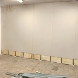 Montaż płyt gipsowo-kartonowych na ścianie z widoczną izolacją z wełny mineralnej u dołu, surowy stan wykończenia, widoczne profile metalowe na podłodze.