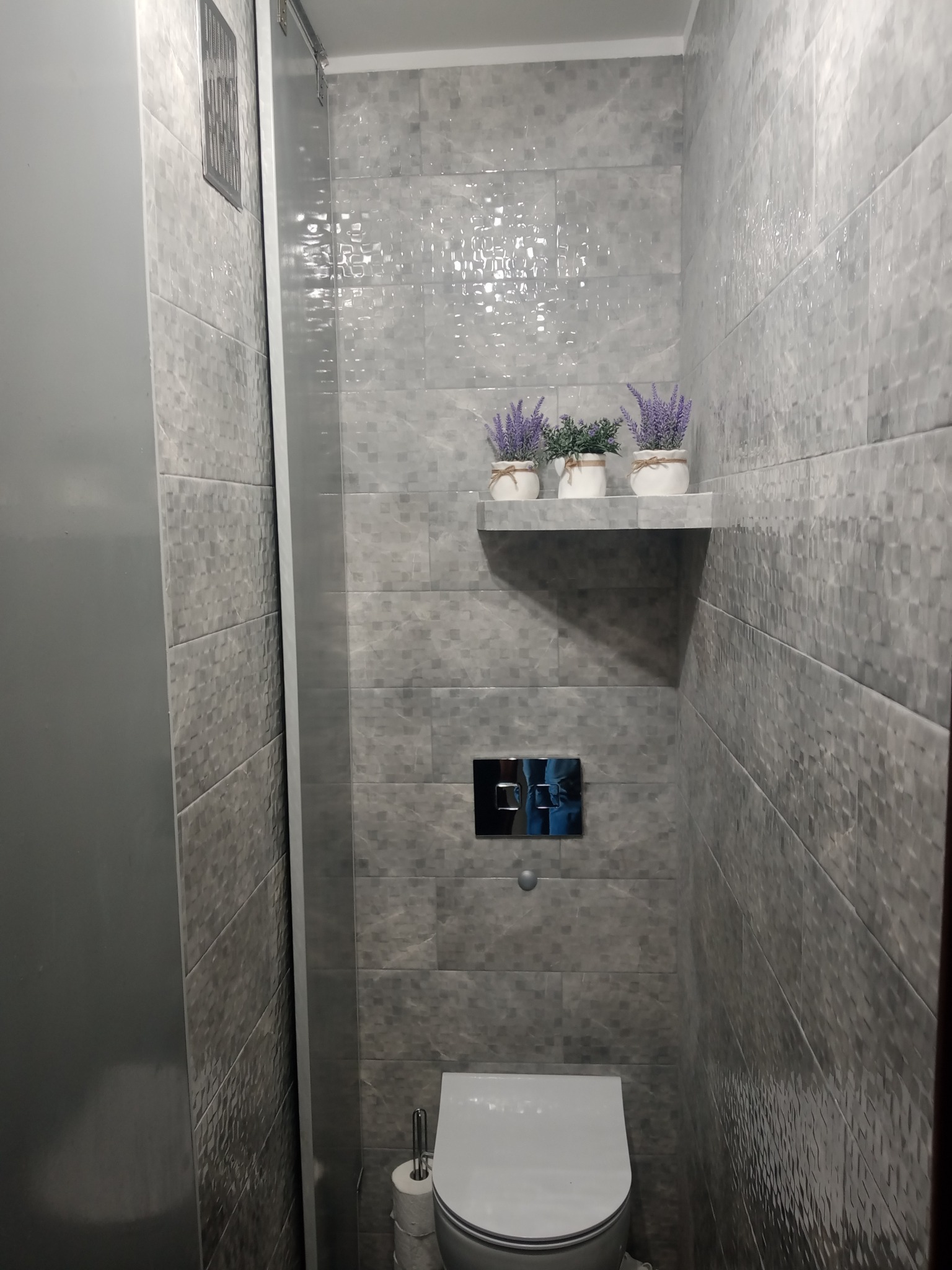 Wąska łazienka wykończona szarymi, błyszczącymi płytkami z delikatnym wzorem, z podtynkową spłuczką, toaletą, półką z trzema doniczkami z lawendą i rolką papieru toaletowego po lewej stronie.