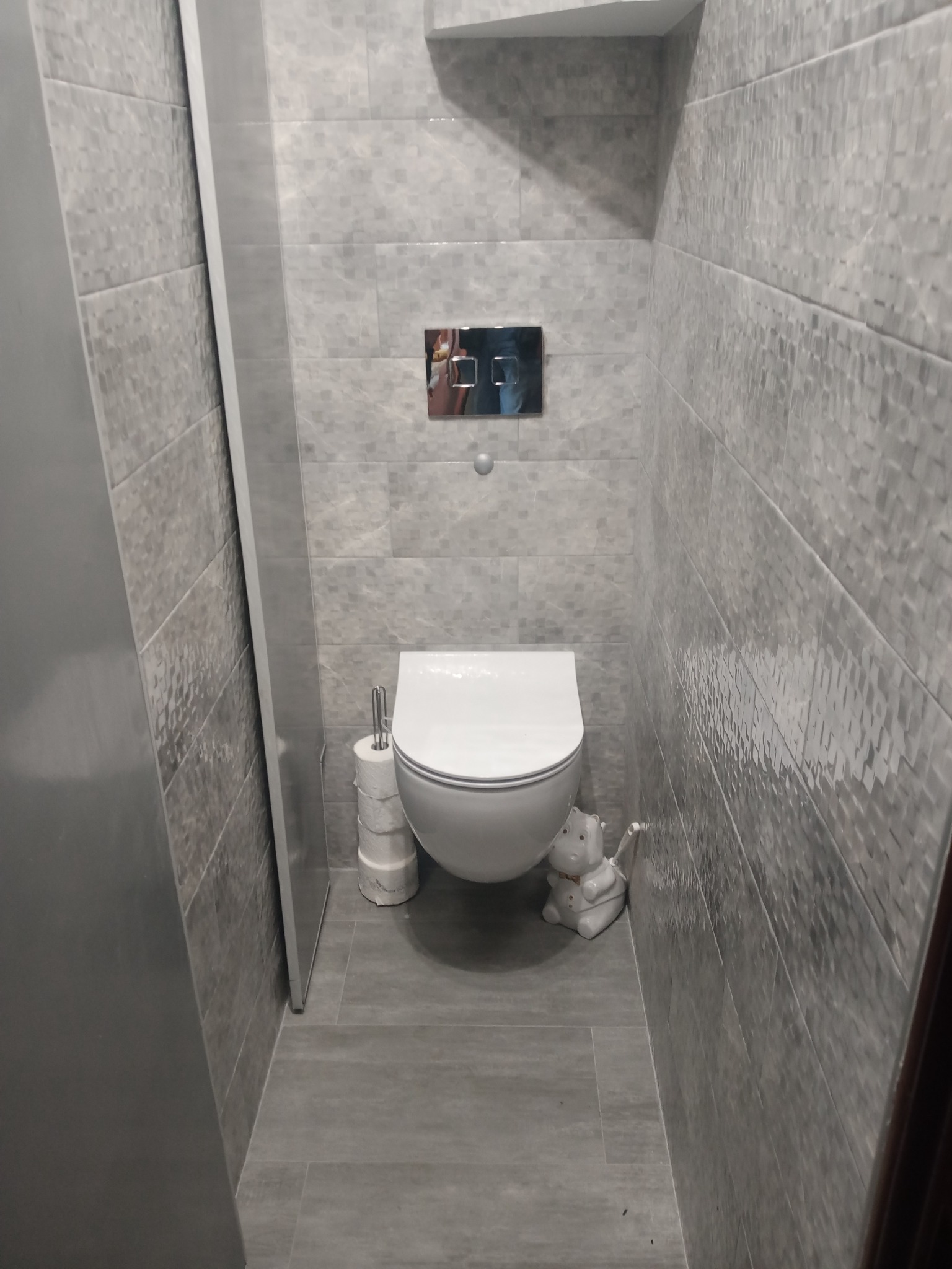 Nowoczesna toaleta w szarej łazience z dekoracyjnymi płytkami, wisząca muszla klozetowa, uchwyt na papier toaletowy i ceramiczna figurka hipopotama.