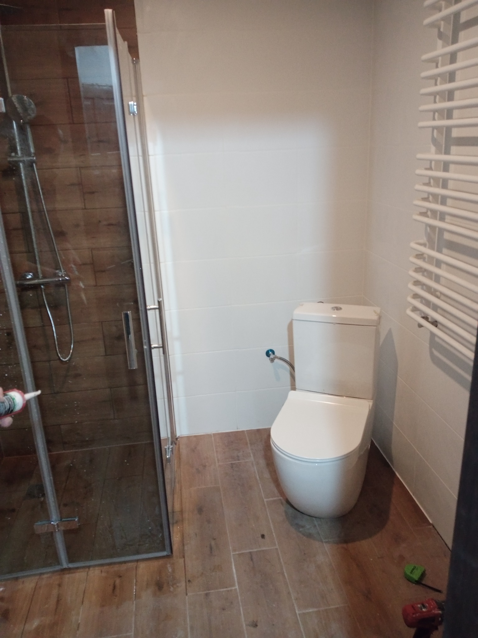 Montaż kabiny prysznicowej z brązowymi płytkami imitującymi drewno, widoczny aplikator silikonu, biała toaleta i grzejnik drabinkowy w wykończonej łazience z brązowymi płytkami podłogowymi.