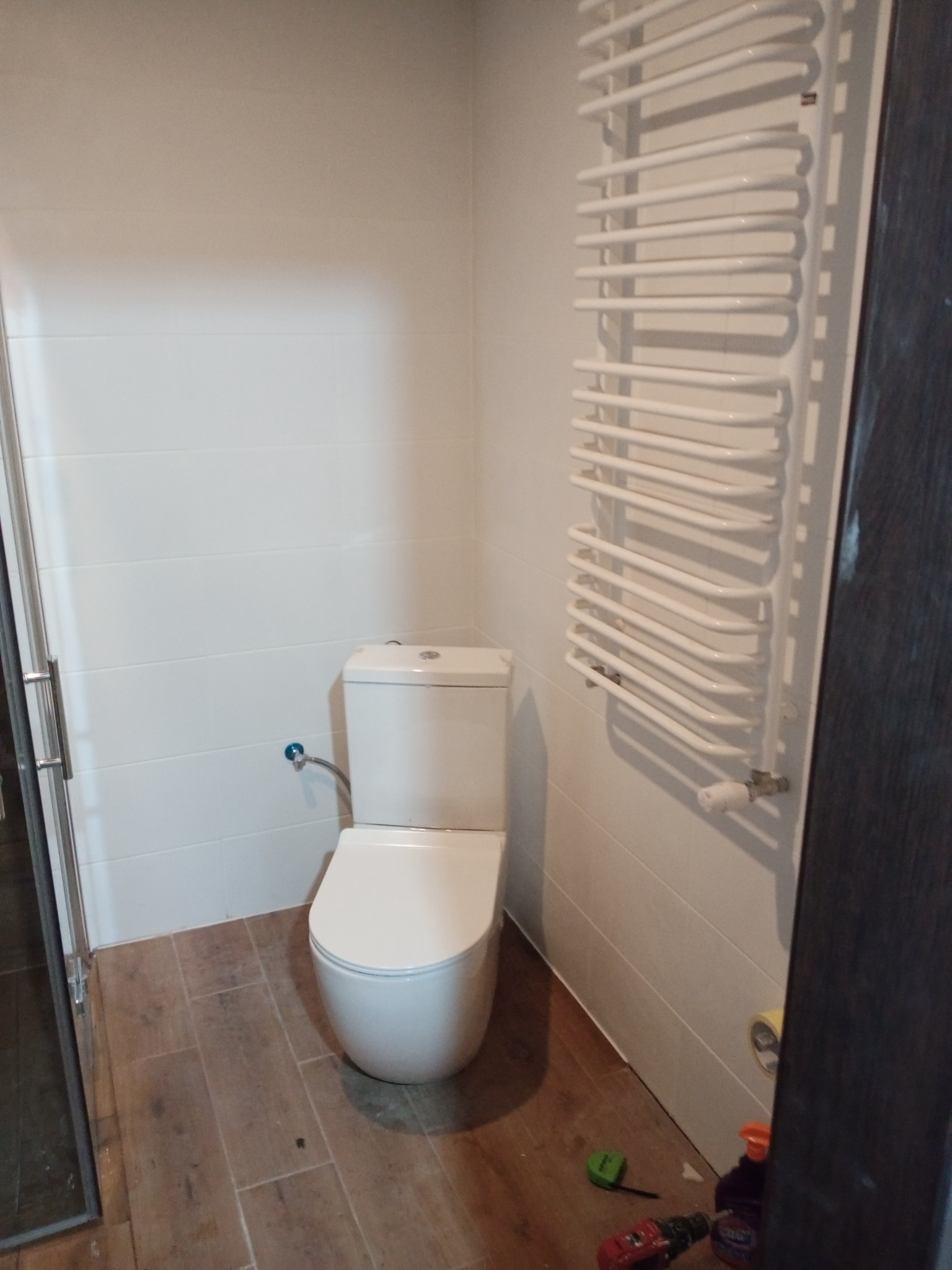 Nowoczesna toaleta w łazience z białymi płytkami i grzejnikiem drabinkowym, widoczne narzędzia i materiały budowlane na podłodze.