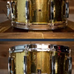 Mosiężny werbel z młotkowaną powierzchnią i chromowanymi obręczami, umieszczony na drewnianej powierzchni z widoczną etykietą 'PURE DUM'.