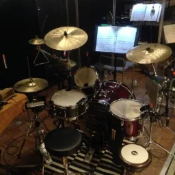 Pełny zestaw perkusyjny z nutami na statywach w tle, widok z góry, z tamburynem i elektronicznym padem perkusyjnym po lewej stronie.