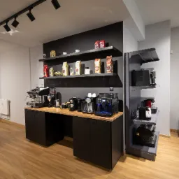 Wnętrze biura lub sklepu z ekspresami do kawy, prezentacja różnych modeli ekspresów na blacie i półkach, obok widoczny fotel i doniczka z rośliną.