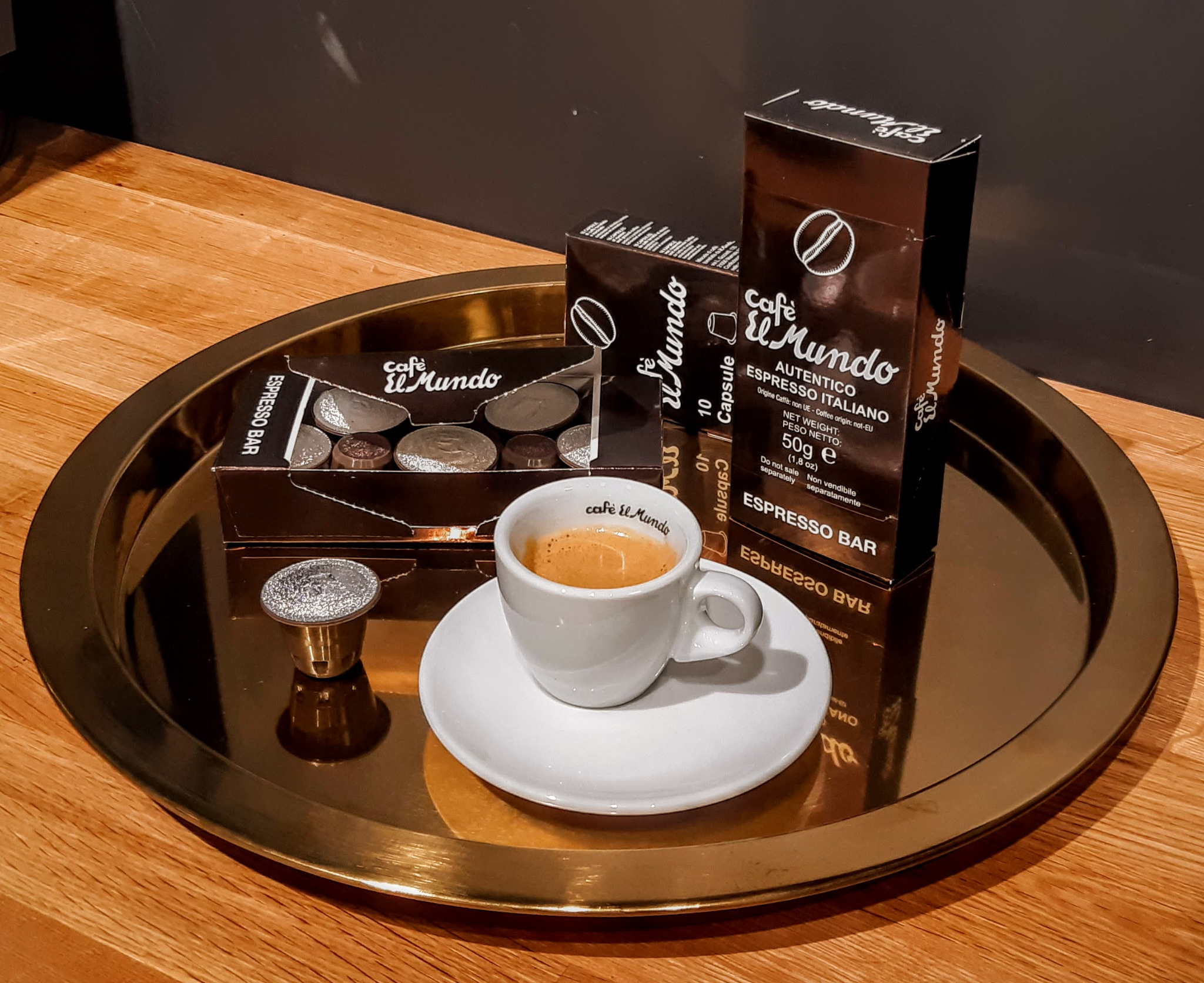 Filiżanka espresso z logo Cafe El Mundo na białym spodku, obok kapsułka i opakowania z kapsułkami na złotej tacy.