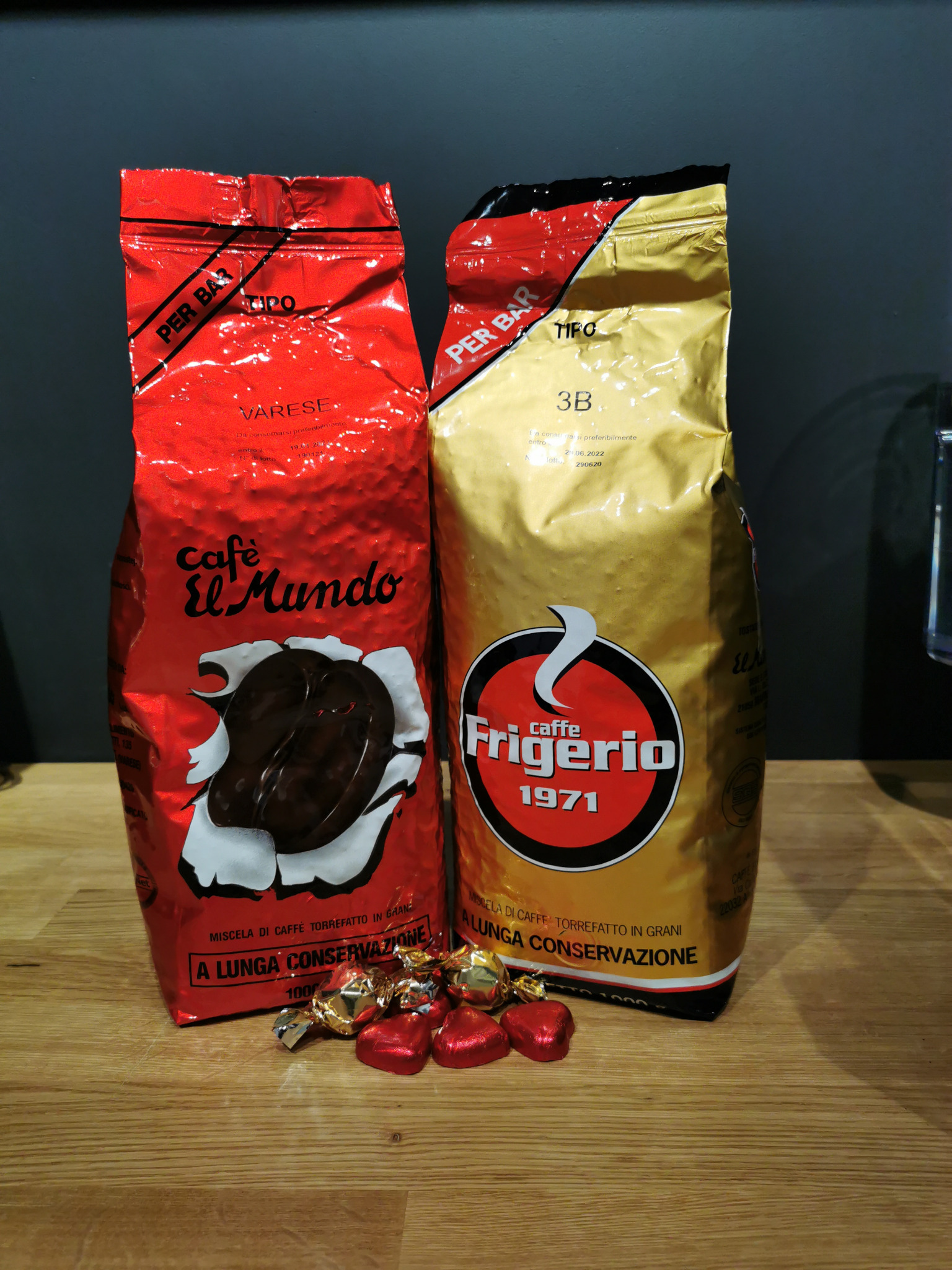 Dwa opakowania kawy ziarnistej Cafe El Mundo i Caffe Frigerio obok siebie, z kilkoma czerwonymi cukierkami w kształcie serca u podstawy, na drewnianym blacie.